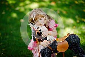 Little preschool girl hugging with rocking horse toy. Happy child in princess dress on sunny summer day in garden. Girl