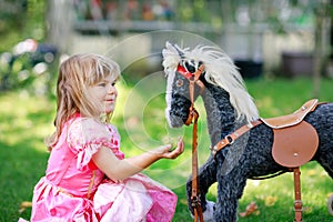 Little preschool girl hugging with rocking horse toy. Happy child in princess dress on sunny summer day in garden. Girl