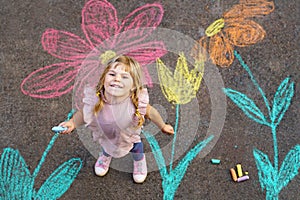 Little preschool girl painting with colorful chalks flowers on ground on backyard. Positive happy toddler child