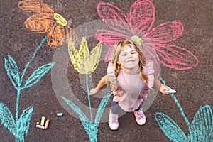 Little preschool girl painting with colorful chalks flowers on ground on backyard. Positive happy toddler child