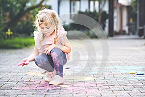 Little preschool girl painting with colorful chalks flowers on ground on backyard. Positive happy toddler child drawing