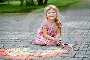 Little preschool girl painting rainbow with colorful chalks on ground on backyard. Positive happy toddler child drawing