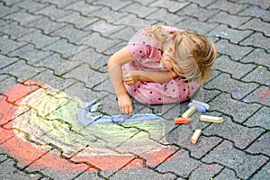 Little preschool girl painting rainbow with colorful chalks on ground on backyard. Positive happy toddler child drawing