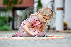 Little preschool girl painting rainbow with colorful chalks on ground on backyard. Positive happy toddler child drawing