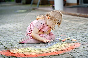 Little preschool girl painting rainbow with colorful chalks on ground on backyard. Positive happy toddler child drawing