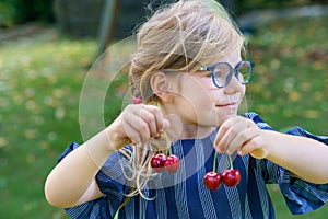Little preschool girl picking and eating ripe cherries from tree in garden. Happy child with glasses holding fresh