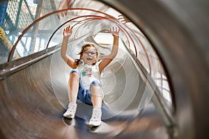 Little preschool girl riding from childrens slides on playground. Happy smiling child having fun in game center for kids