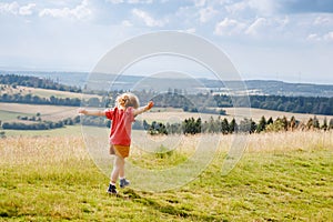 Little preschool girl running and walking through meadow with blooming field flowers, with view on hills and mountains
