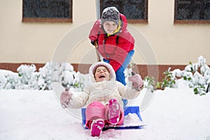 Little preschool girl and school boy playing with snow in winter. Brother sliding cute sister on shovel. Happy smiling