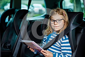 Little Preschool Girl Sitting in Her Car Seat. Happy Child with Eyeglasses Reading a Book, Smiling on the Way to Family