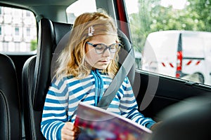 Little Preschool Girl Sitting in Her Car Seat. Happy Child with Eyeglasses Reading a Book, Smiling on the Way to Family