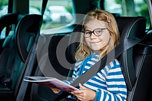 Little Preschool Girl Sitting in Her Car Seat. Happy Child with Eyeglasses Reading a Book, Smiling on the Way to Family