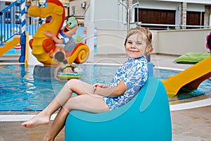 Little preschool girl sitting by outdoor swimming pool in hotel resort. Child learning to swim in outdoor pool