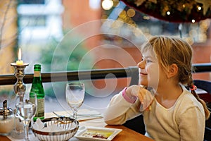 Little preschool girl sitting in restaurant near window decorated for Christmas. Child waiting for food. Happy family