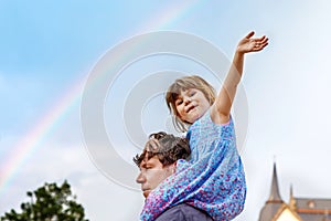 Little preschool girl sitting on shoulder of father. Happy toddler child and man observing rainbow on sky after summer
