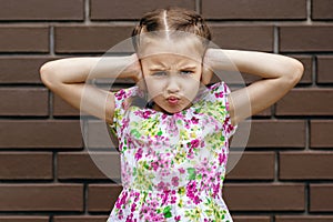 A little preschool girl stands by a brick wall, covers her ears with her hands and is very unhappy