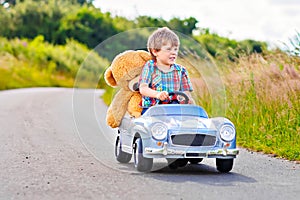 Little preschool kid boy driving big toy car and having fun with playing with his plush toy bear, outdoors. Child