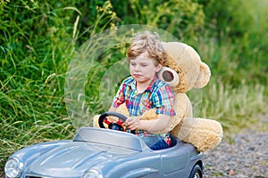 Little kid boy driving big toy car with a bear, outdoors.