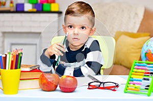 Little preschooler boy is thoughtful and sits at a desk with a book, a globe, a notebook and pencils.