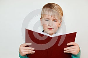 A little pretty boy with blond hair and blue eyes holding red opened book reading interesting stories. Seven years old boy reading