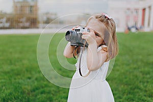 Little pretty child baby girl in light dress take picture on retro vintage photo camera on green grass in park. Mother