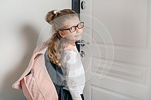 Little pretty girl 6, 7 years old with school backpack. Smiling girl standing near the front door of the house, child goes to