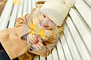 Little pretty girl in a beige coat sitting on a bench and laughing in the park in autumn