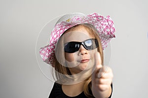 little pretty girl in black sunglasses and a beach hat smiles and raises her thumb up