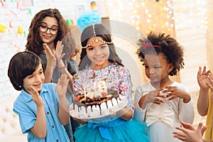 Little pretty girl with pendant chain on her head holds cake with candles at celebration of birthday.