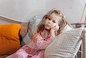 A little pretty girl rests on a pillow on a bed in a children`s bedroom. Thoughtfulness and dreaminess