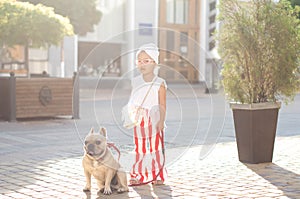 little pretty girl stylishly dressed with striped red-white pants walking her French bulldog dog