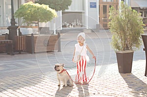 little pretty girl stylishly dressed with striped red-white pants walking her French bulldog dog