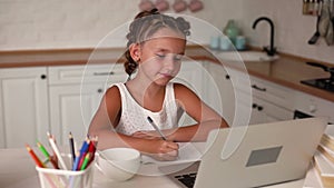 Little pretty girl with two ponytails sitting at kitchen at table with exercise book and laptop doing her school lessons because o