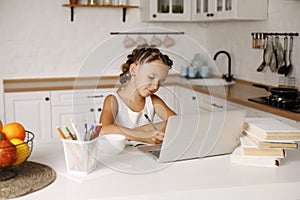 Little pretty girl with two ponytails sitting at kitchen at table with exercise book and laptop doing her school lessons because o