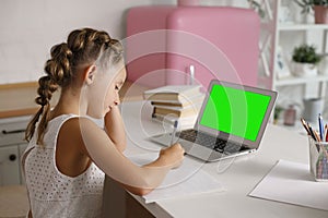 Little pretty girl with two ponytails sitting at kitchen at table with exercise book and laptop doing her school lessons because o