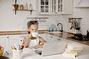 Little pretty girl with two ponytails sitting at kitchen at table with exercise book and laptop drinking tea and doing her school