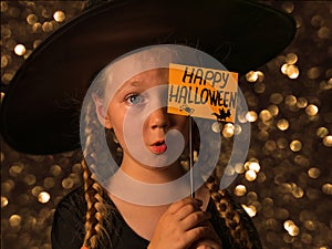 Little pretty girl witch in black wizard hat holds greeting Happy Halloween card says Boo and laughing.Autumn kids party