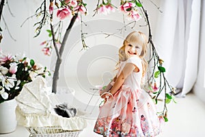 Little pretty smiling blonde 3 years old girl in pink dress rolling white baby carriage with gray and white furry rabbit inside.