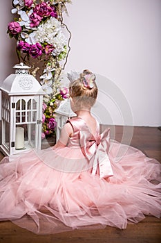 The little princess in a beautiful pink dress sits on the floor near the flower stand and opens the lantern, view from the back.