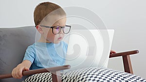 little pupil in glasses sitting in armchair watching opened laptop computer home