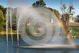 Little rainbow from the water fountain lake, city park pond, water jet beats up against the background of green trees