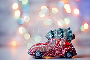 Little red car toy carrying Christmas tree in snow and bokeh christmas lights