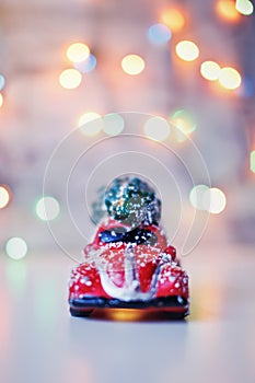 Little red car toy carrying Christmas tree in snow and bokeh christmas lights