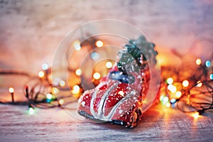 Little red car toy carrying Christmas tree in snow and bokeh christmas lights