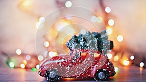 Little red car toy carrying Christmas tree in snow and bokeh christmas lights