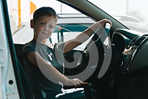 Little red-haired boy sits in front of the car holding steering wheel. Boy looking at the camera and smiling