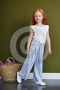 Little red-haired girl with a basket of flowers posing on an olive background. Spring portrait of a redhead girl with blue eyes.