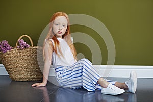 Little red-haired girl with a basket of flowers posing on an olive background. Spring portrait of a redhead girl with blue eyes.