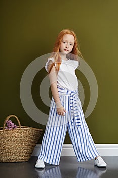 Little red-haired girl with a basket of flowers posing on an olive background. Spring portrait of a redhead girl with blue eyes.