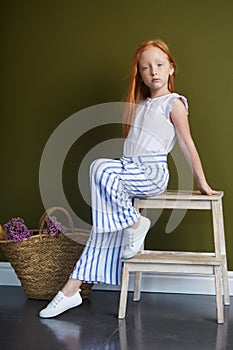 Little red-haired girl with a basket of flowers posing on an olive background. Spring portrait of a redhead girl with blue eyes.